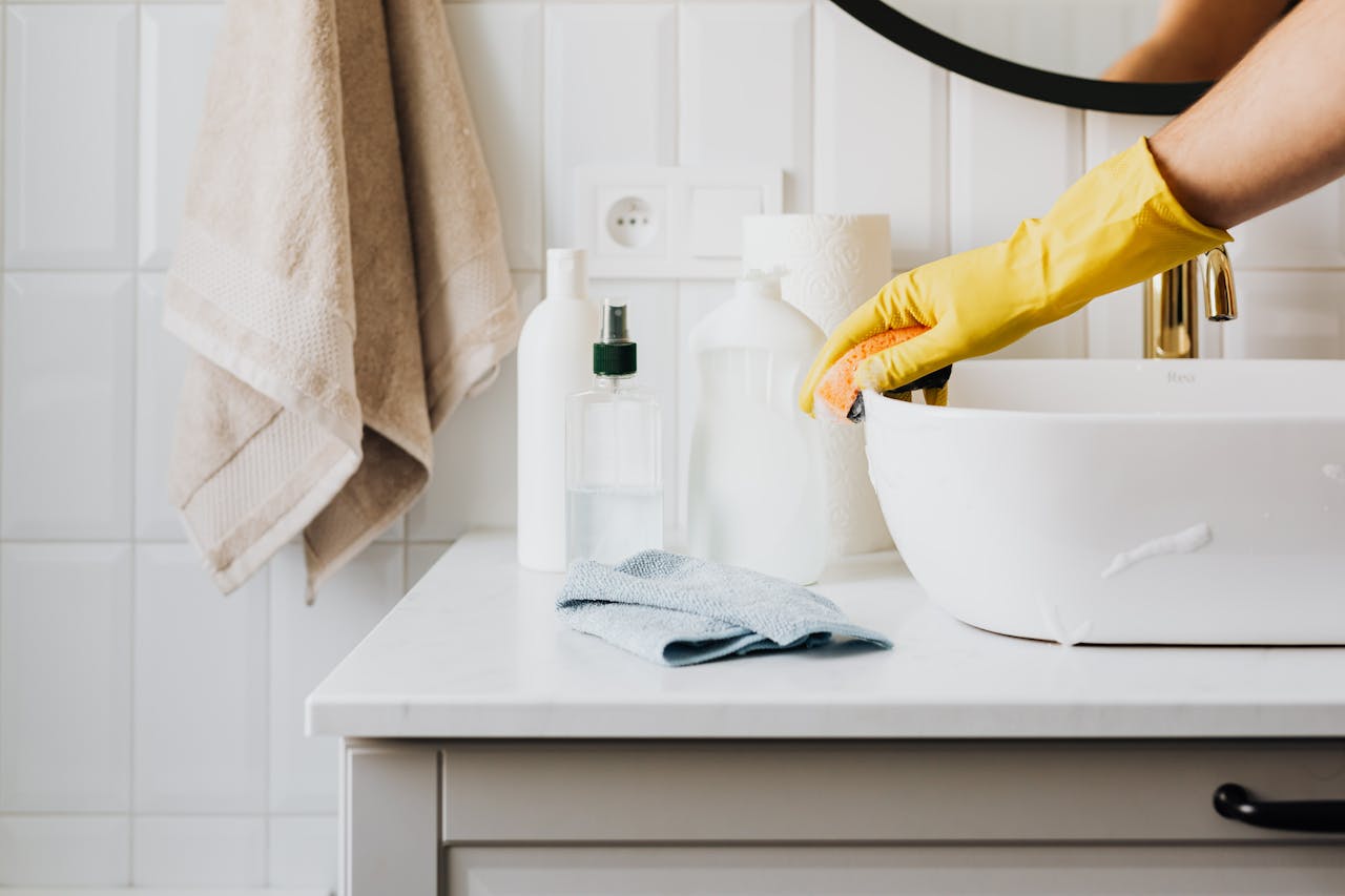 Commercial Cleaning Person cleaning a bathroom sink with yellow gloves and cleaning products on a countertop.